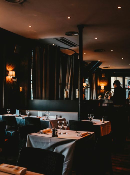 Dimly lit restaurant with set tables, wine glasses, and a person standing near the bar in the background.