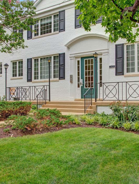 White brick house with black shutters, front steps, iron railing, and well-kept lawn and shrubs.