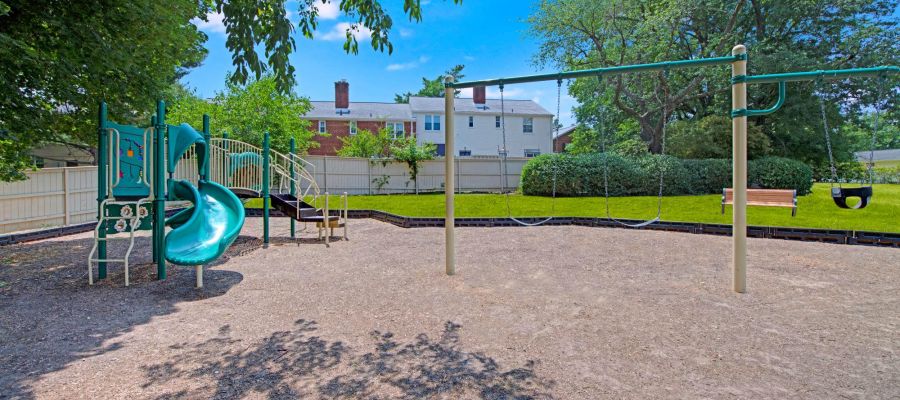 Empty playground with slides, swings, and a bench, surrounded by trees and a white fence on a sunny day.
