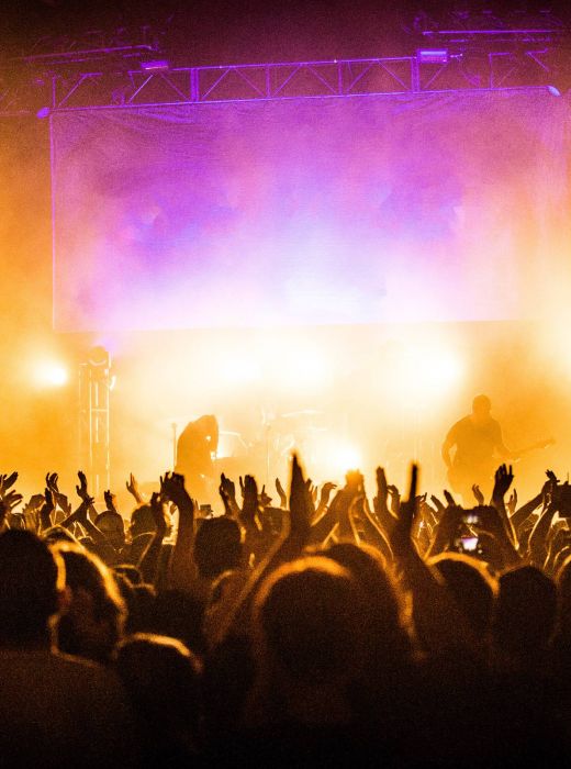 A crowd cheers at a concert with a band playing on stage surrounded by bright, colorful lights.